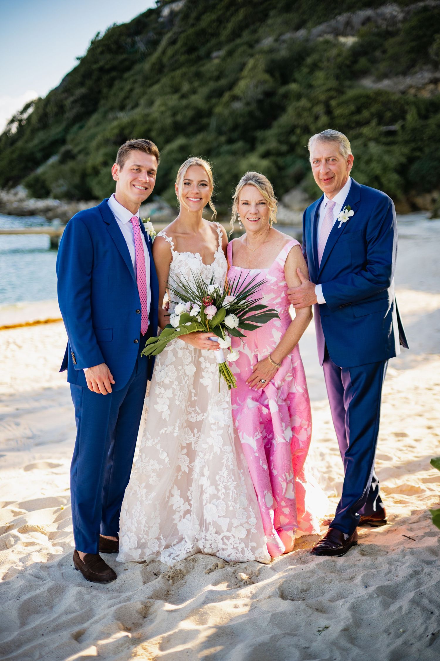 Wedding party poses together on a scenic beach at sunset with blue suits and pink dresses.