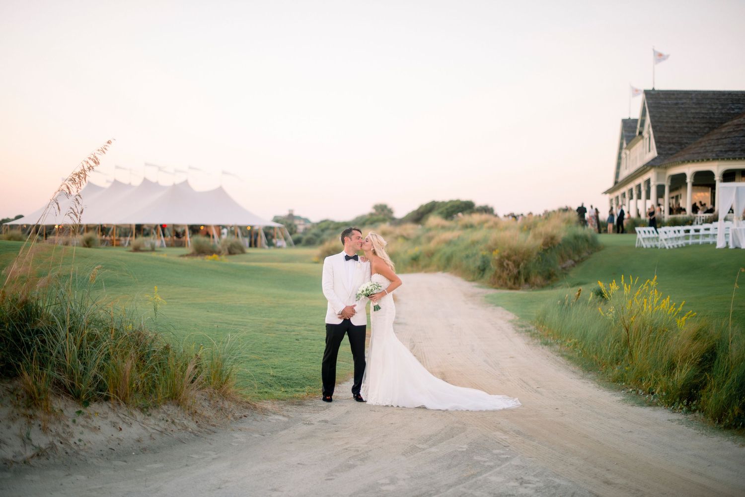 A couple embraces on a sandy path at sunset with a golf course and coastal house in the background.