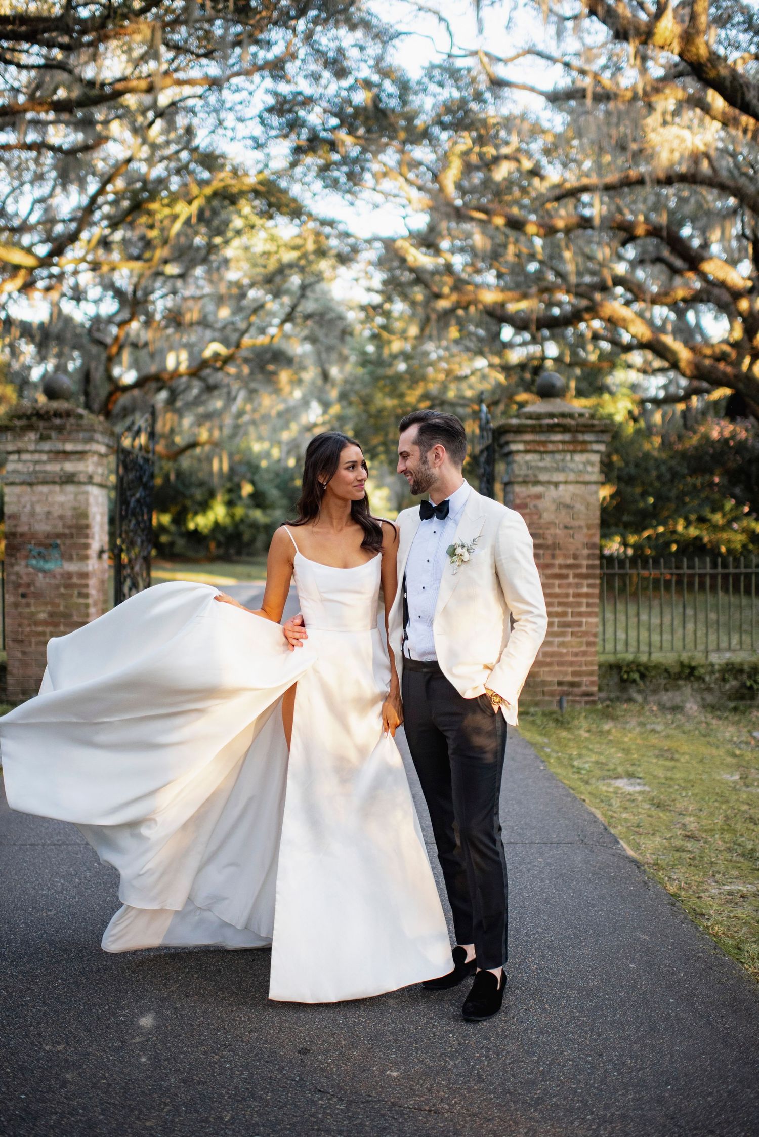 A couple in elegant wedding attire strolls along a tree-lined path at sunset, with the bride's white dress flowing in the breeze.