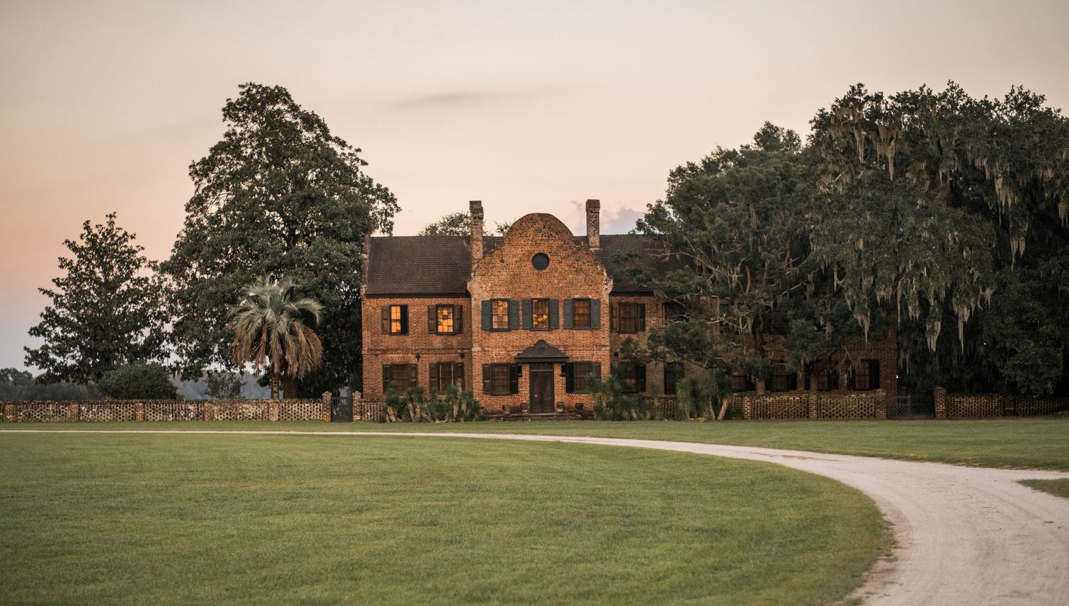A long driveway leads to a historic brick mansion surrounded by green lawns and mature trees.