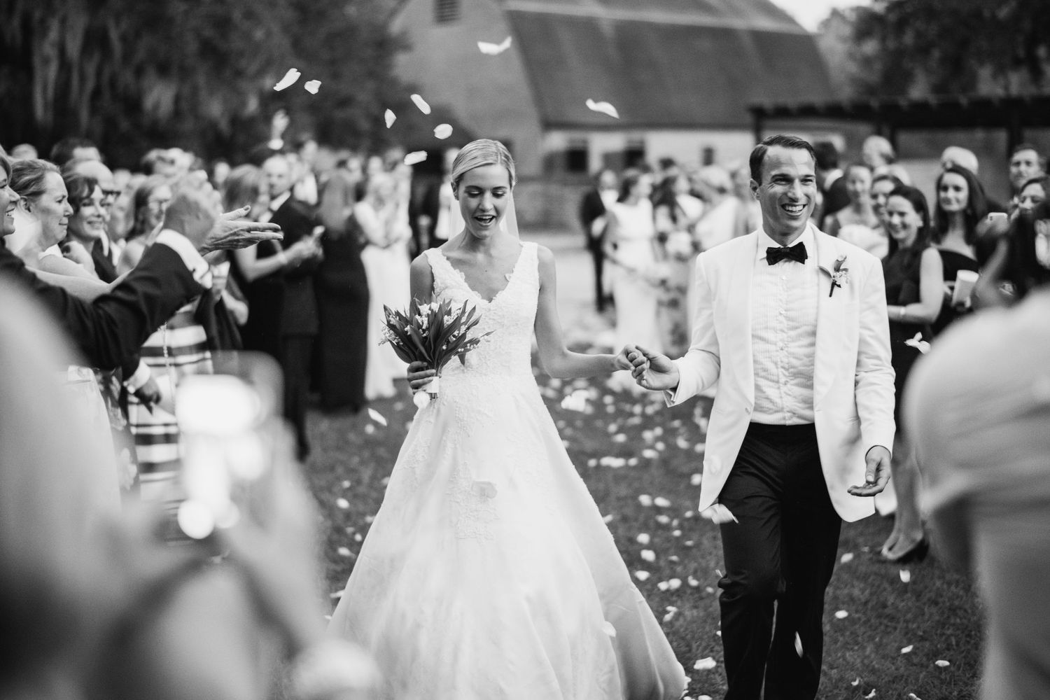 A black and white wedding photo showing newlyweds walking through flower petals being tossed by guests outside.
