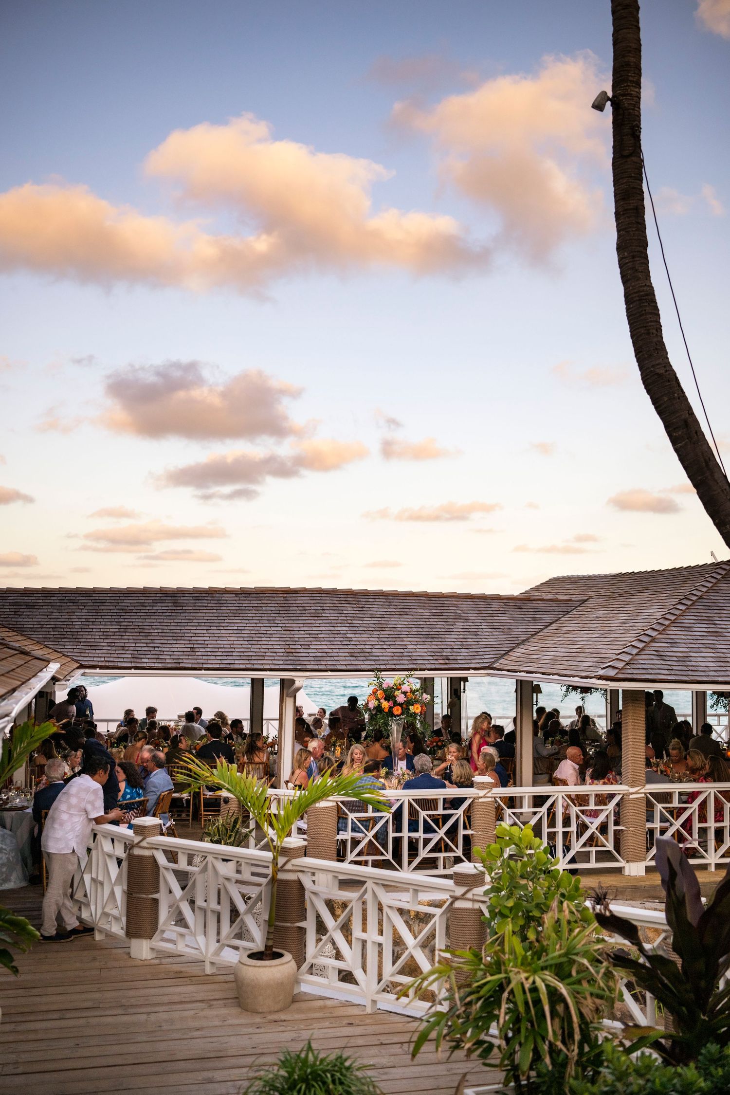 Wedding guests gather on a covered oceanfront deck during sunset.