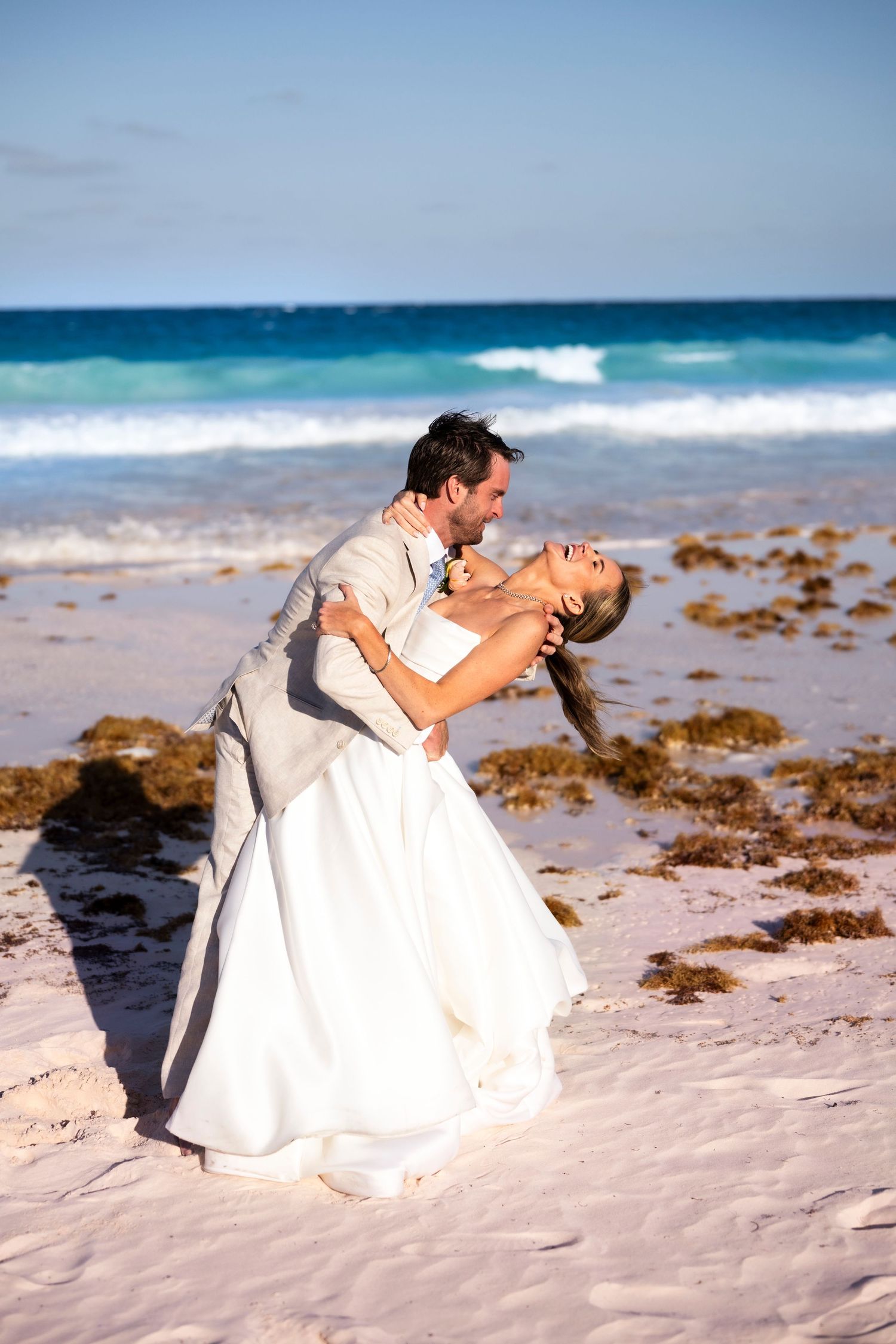 Romantic beach wedding photo with couple dancing in sand by ocean waves.