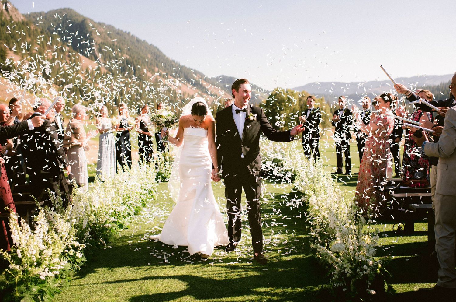 Wedding guests shower newlyweds with flower petals during an outdoor garden celebration on a sunny day.
