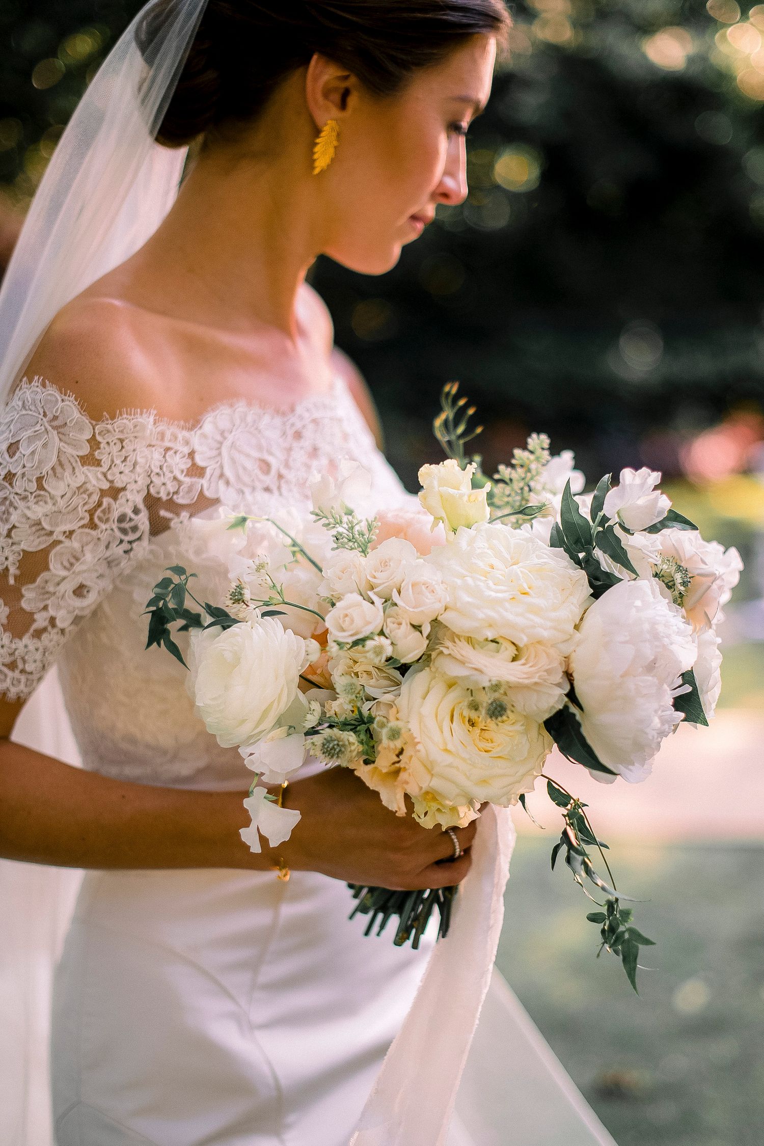Elegant white peony bouquet held against a lace wedding dress in soft natural lighting.