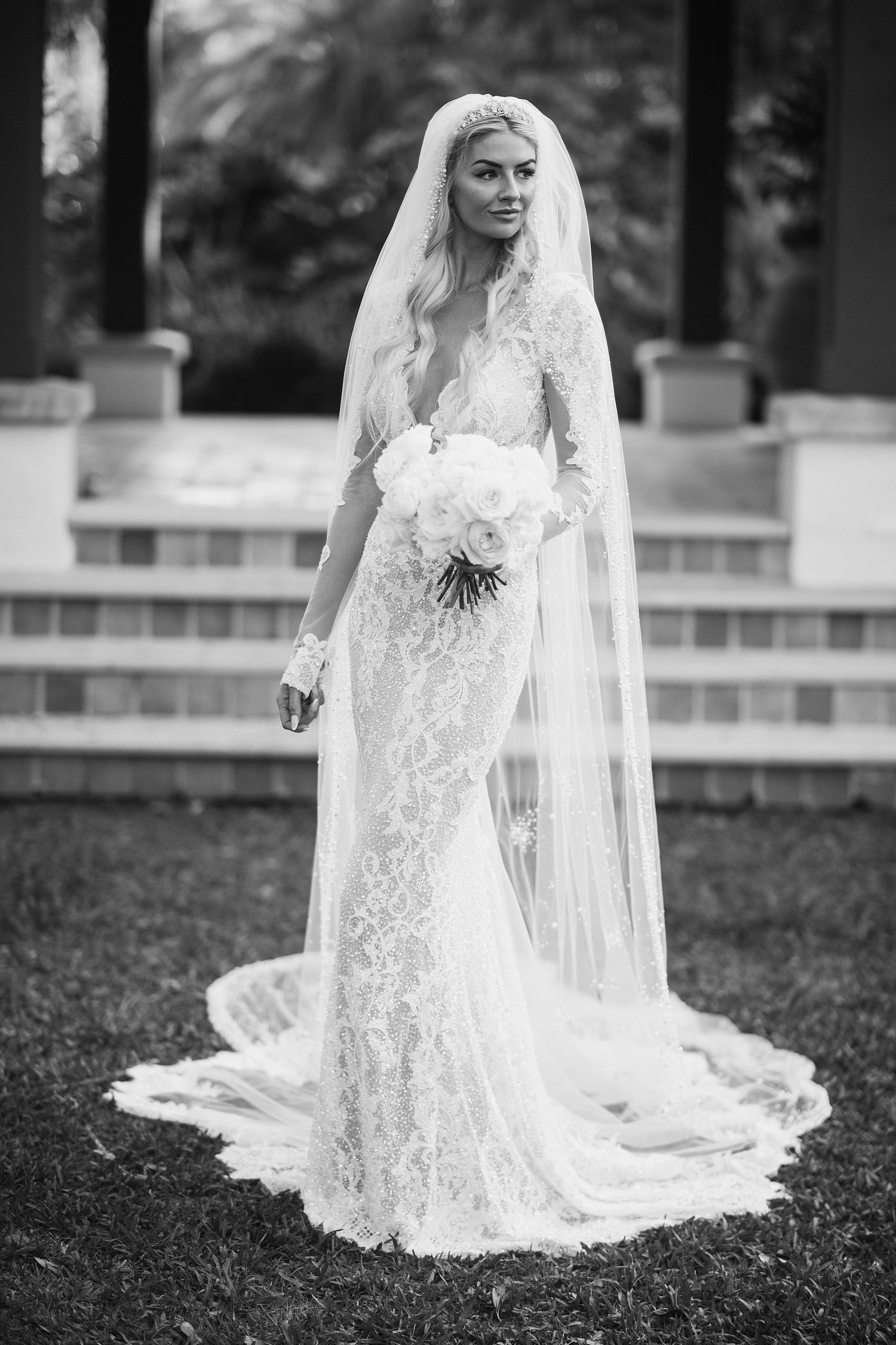 A bride in lace gown and veil poses gracefully on garden steps.