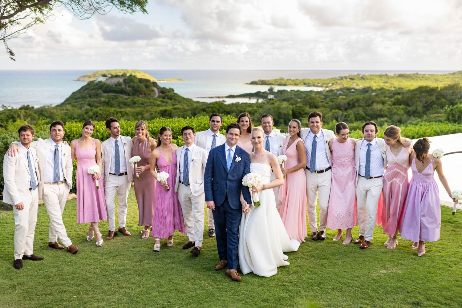 Wedding party in pink dresses and light suits gathered on a scenic hilltop overlooking the ocean.