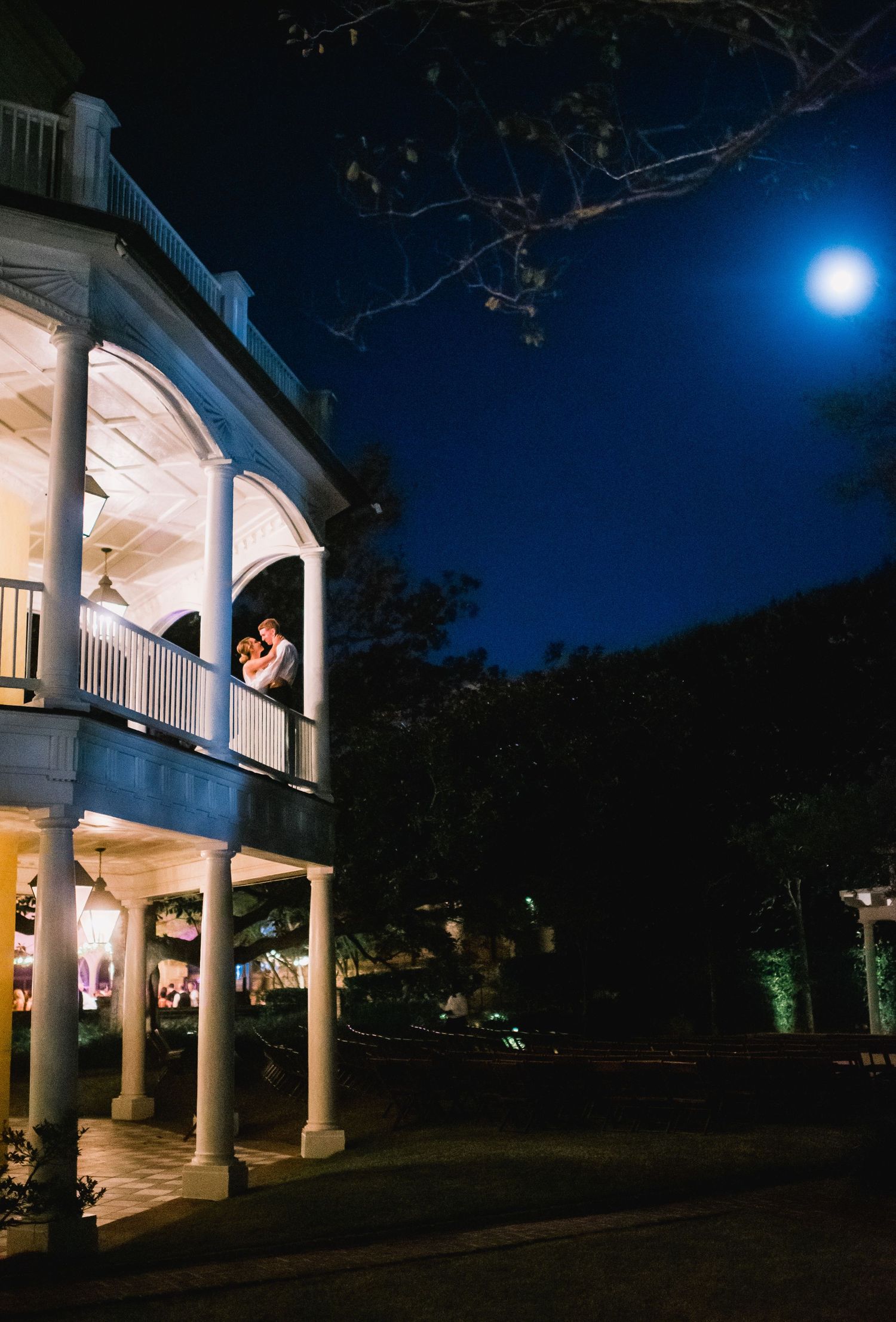 Elegant white mansion with illuminated columns and porches at night under a bright full moon.