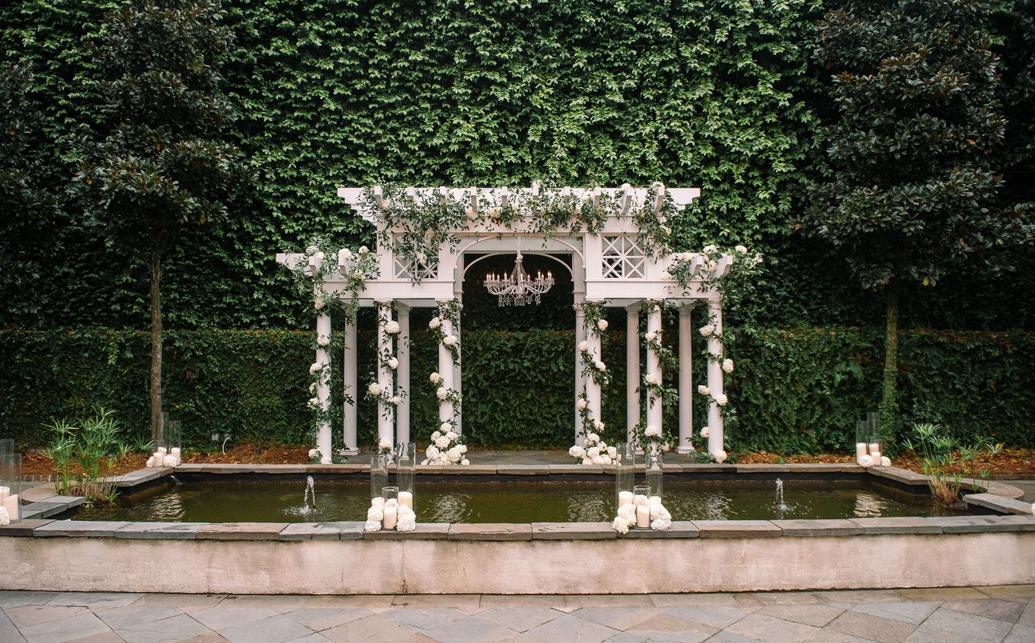 An ornate white stone gazebo with columns and floral decorations stands in front of an ivy-covered wall in a formal garden.