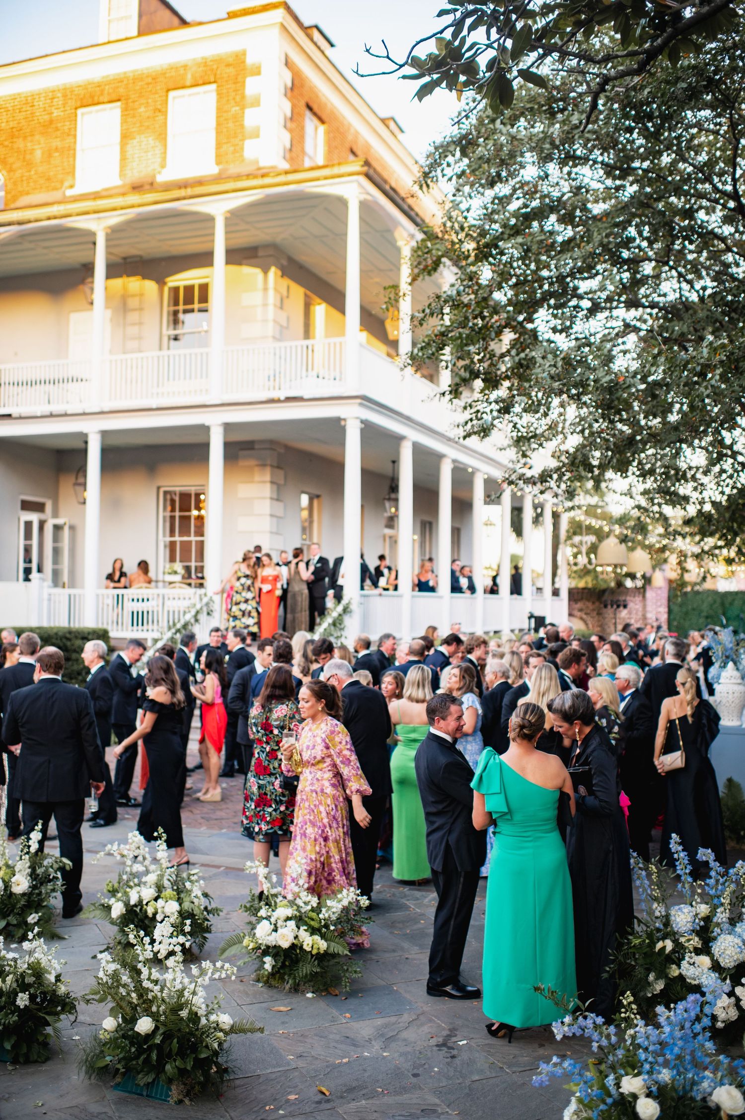 Large group of wedding guests gather outside a white colonial-style mansion during an elegant outdoor reception.
