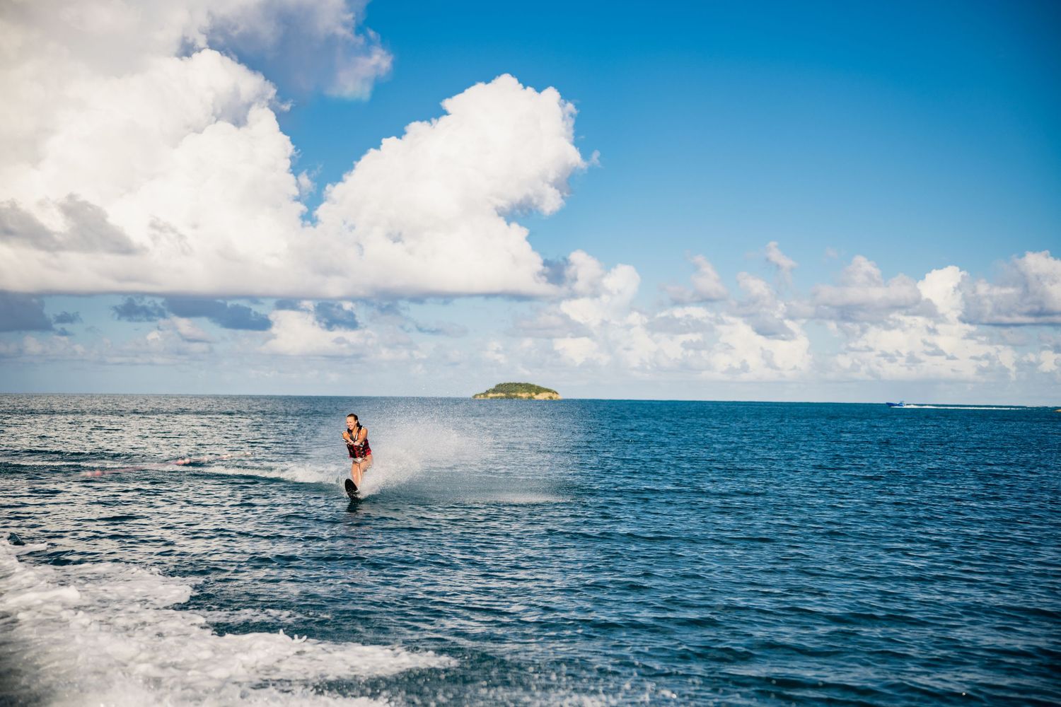 A surfer rides a wave across a vast blue ocean under white puffy clouds and bright sunlight.