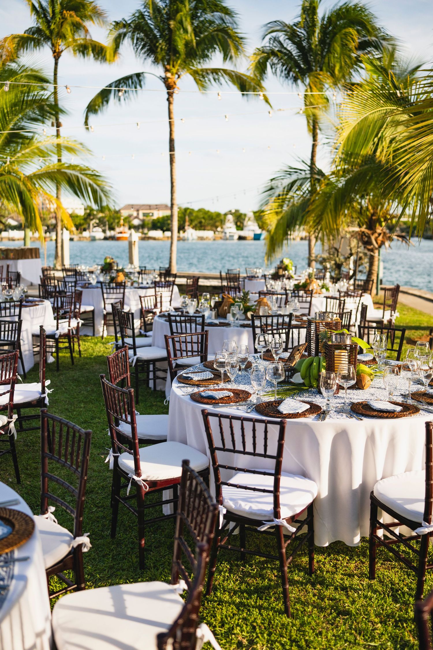 Elegant outdoor wedding reception setup with white-clothed tables and chairs under palm trees by the ocean.