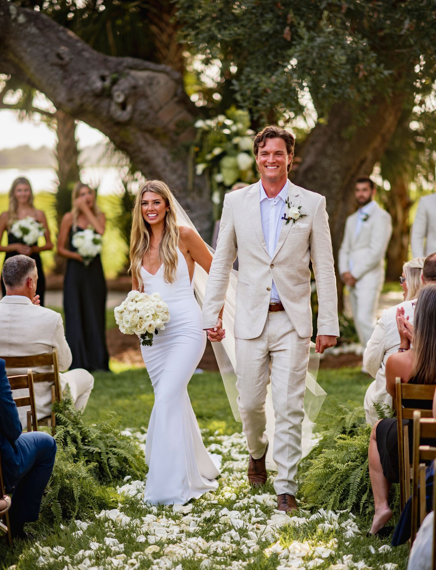Wedding ceremony guests seated outdoors under oak trees with white chairs and floral decorations.