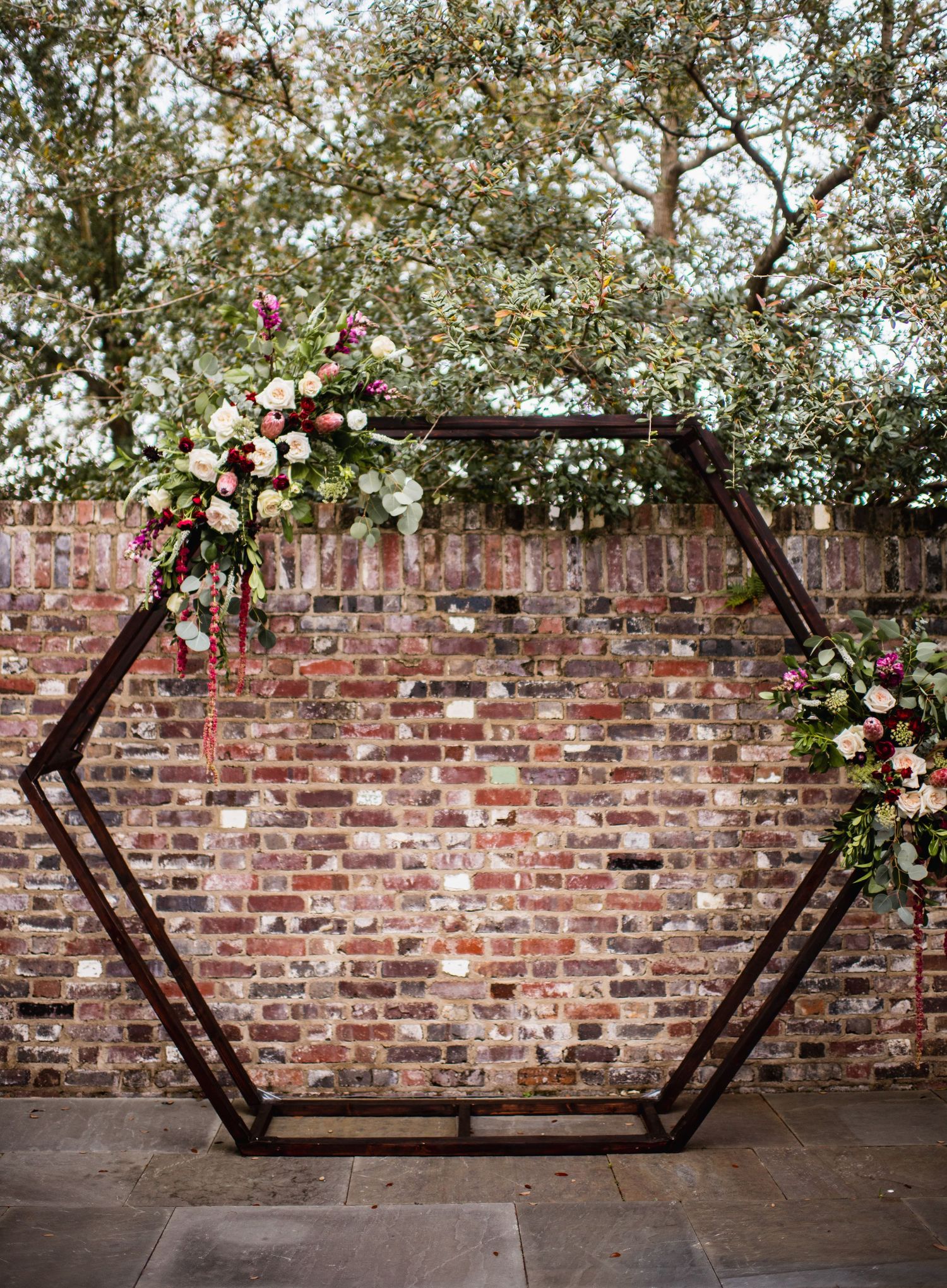 Three wooden hexagonal frames adorned with floral arrangements against a rustic brick wall create an elegant wedding backdrop.