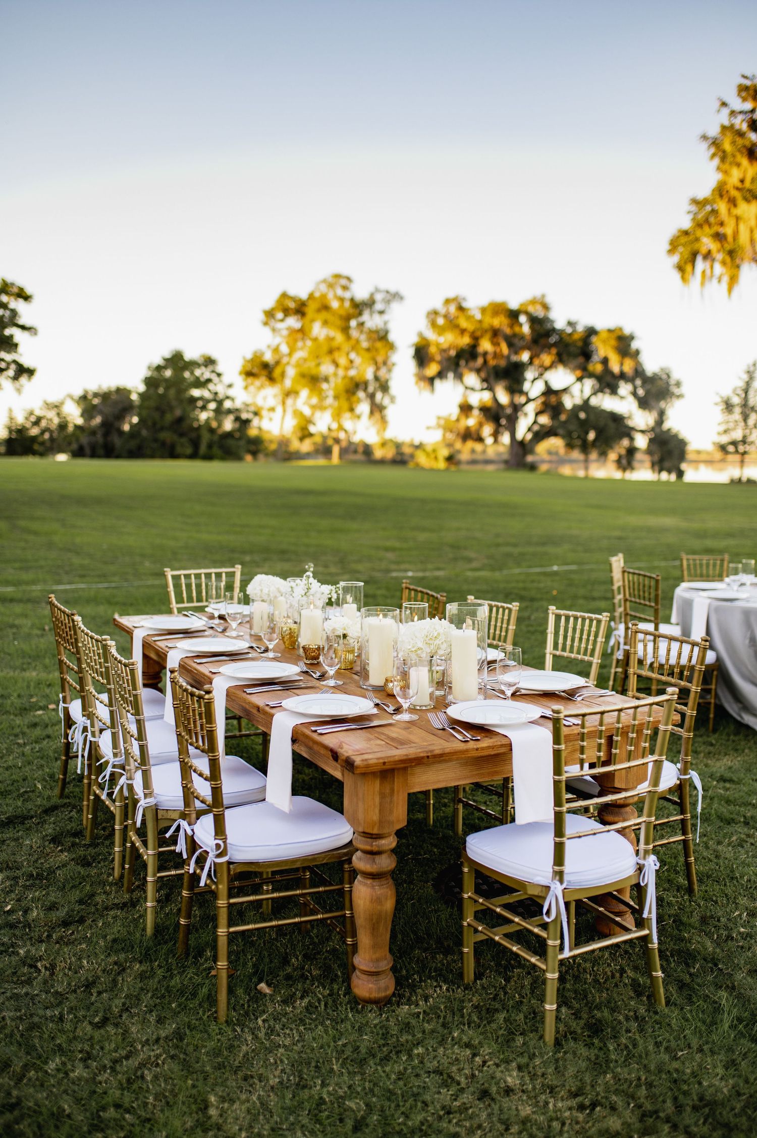 Rustic outdoor wedding reception tables with gold chiavari chairs and wooden farm tables set up on grassy lawn at sunset.