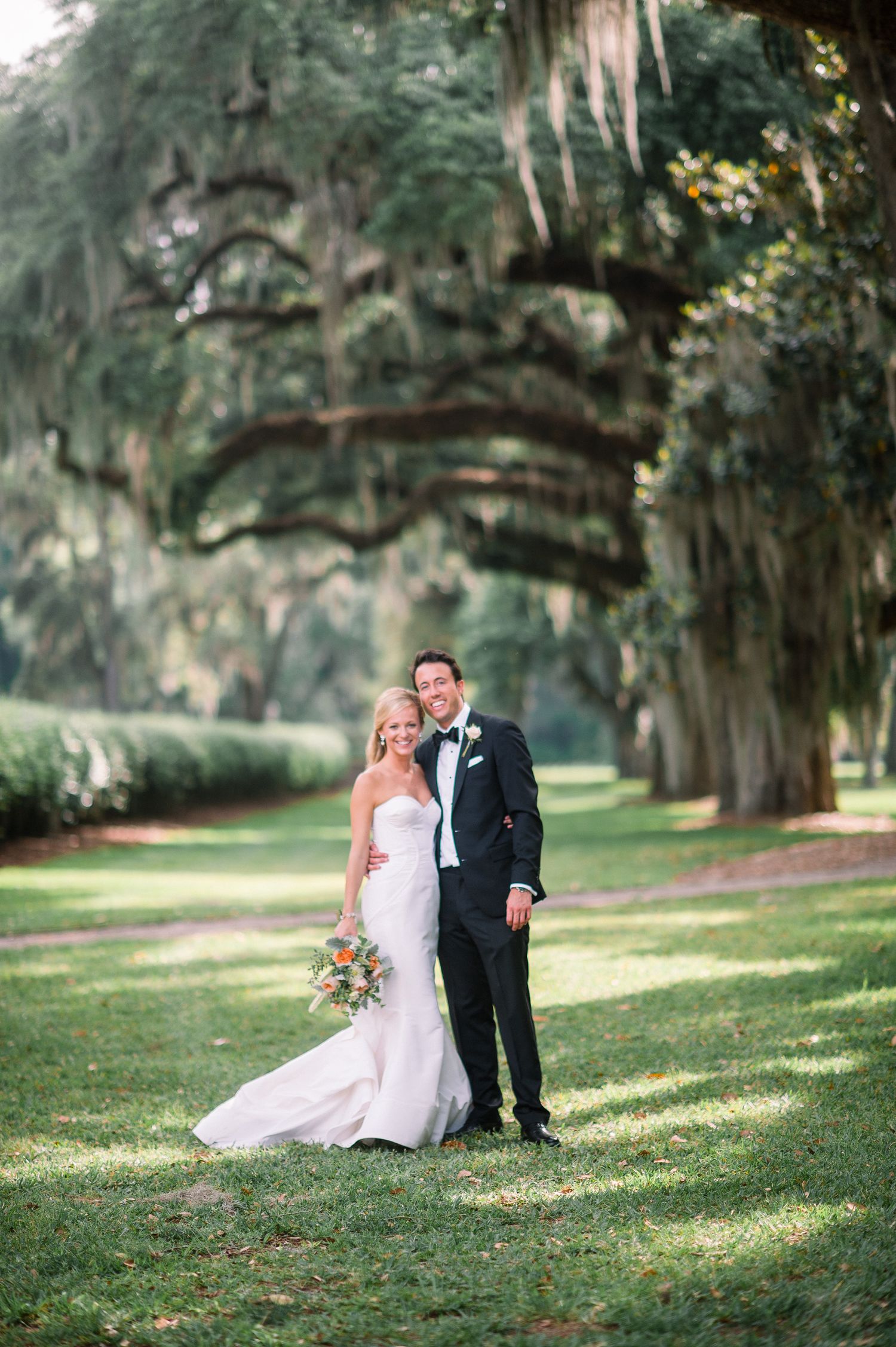 Romantic wedding couple poses together under majestic oak trees draped with Spanish moss at an outdoor venue.
