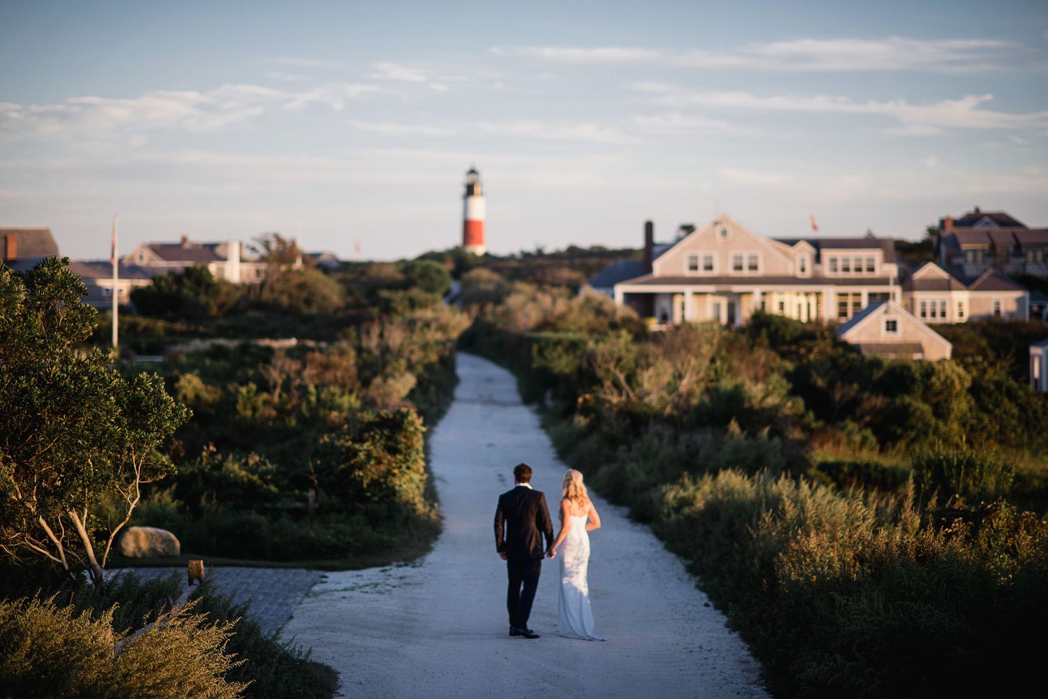 A couple in wedding attire walks along a coastal path with scenic ocean views and beachfront homes in the background.