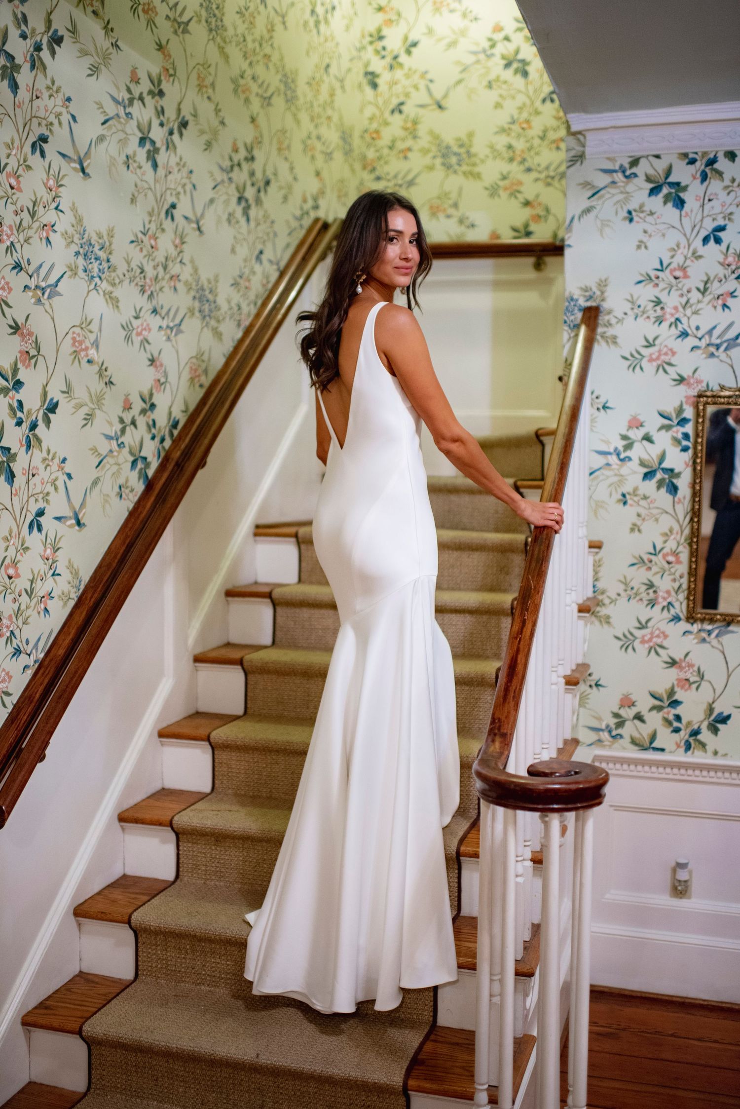 Elegant white wedding dress displayed on wooden staircase with floral wallpaper in background.