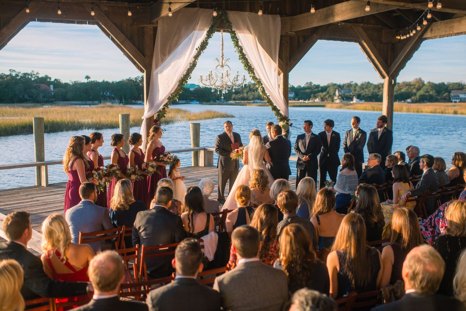 Wedding ceremony taking place under a wooden gazebo overlooking a scenic waterfront with guests seated in rows.