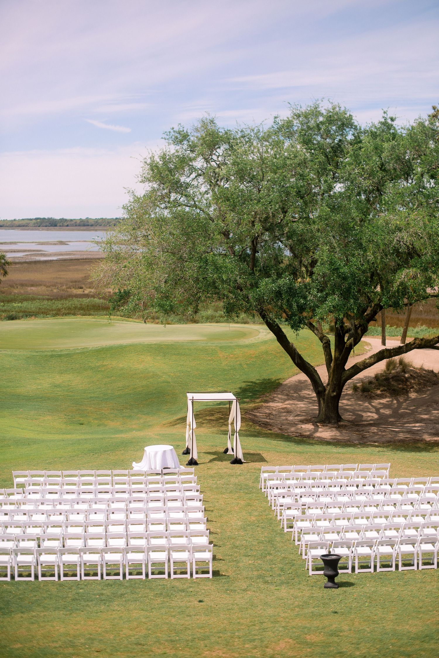 White chairs arranged for an outdoor wedding ceremony under olive trees beside a scenic waterfront landscape.