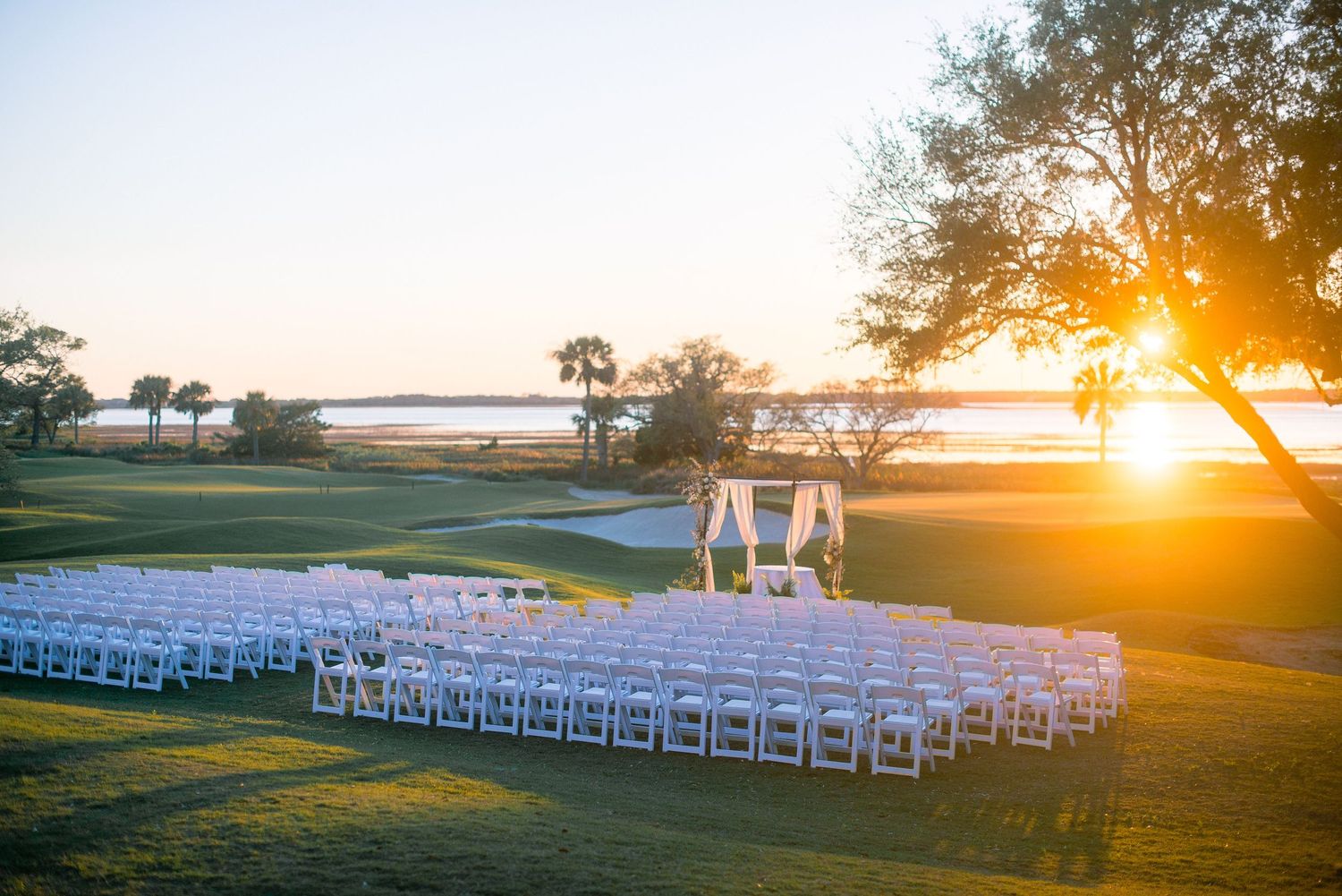 White wedding chairs arranged on a golf course during a golden sunset ceremony with ocean views in the background.