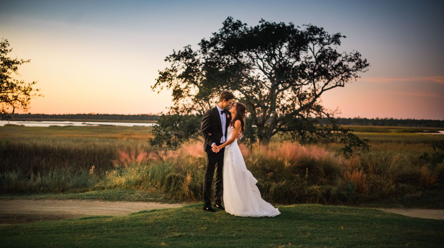 A couple embraces during golden hour sunset with a tree silhouette and marsh landscape in the background.