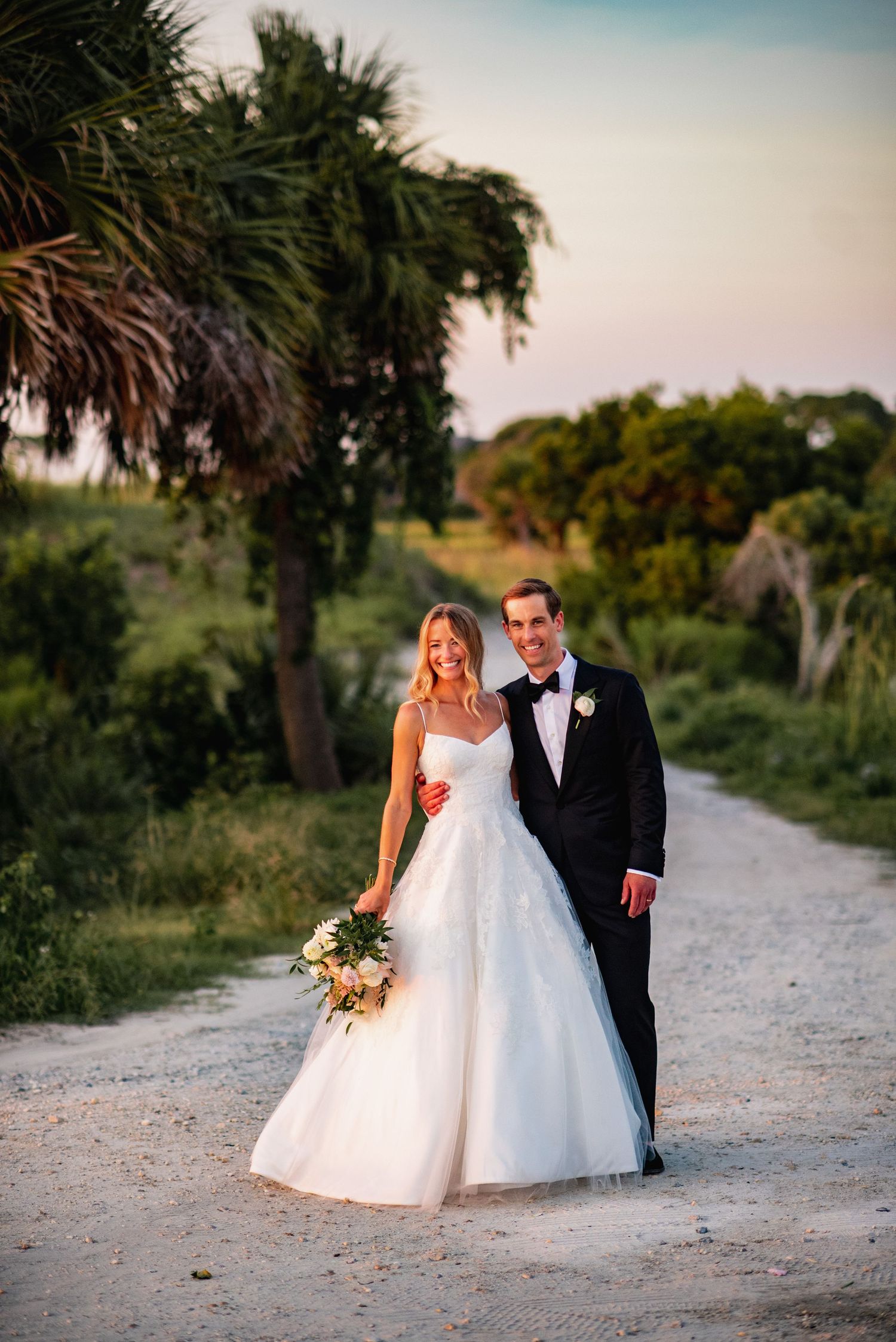 A couple in wedding attire walks along a palm-lined sandy path at sunset during their beach wedding photoshoot.