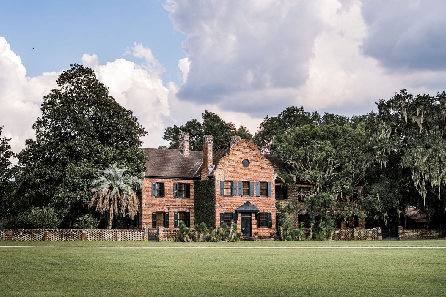 A panoramic view of a red brick mansion with Spanish moss draped oak trees on a sprawling green lawn estate.