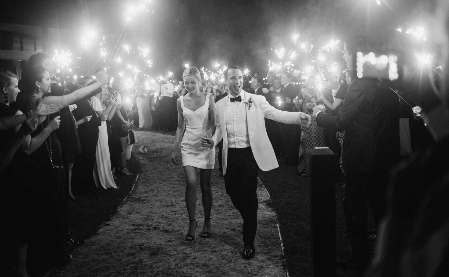 A joyous couple exits their wedding celebration through a tunnel of guests holding sparklers in black and white.
