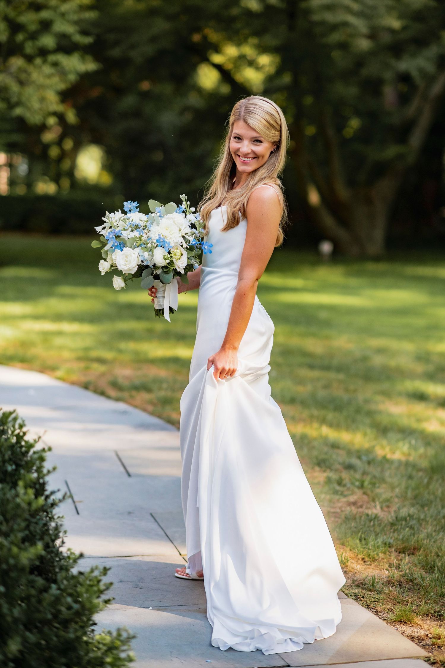 A bride in a flowing white wedding dress holds a blue and white floral bouquet while standing on a garden path.