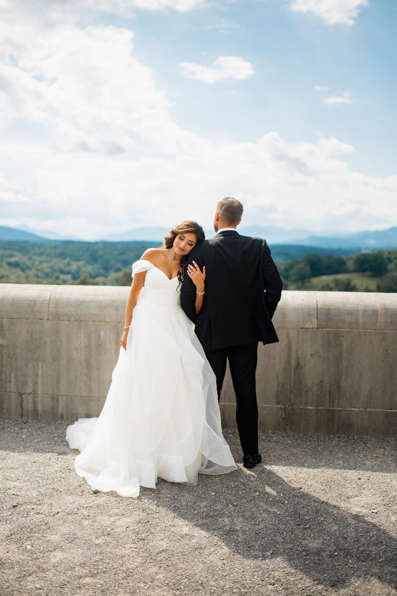 A couple in wedding attire stands on a concrete overlook with mountain views in the background.