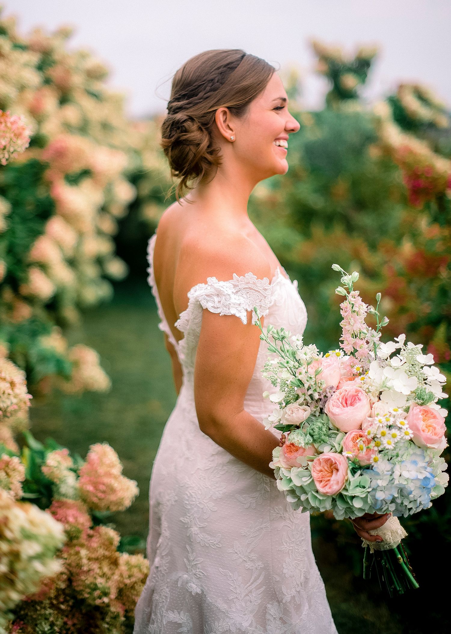 A bride in a lace wedding dress holds a pastel bouquet while posing in a garden setting.