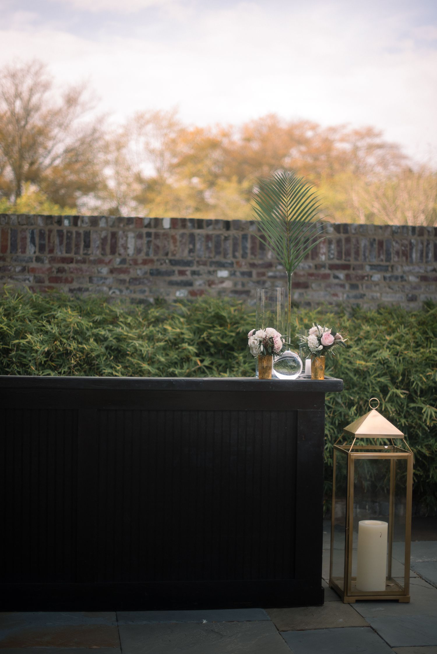A decorative gold lantern and floral arrangement sit atop a black surface against a stone wall with autumn trees in background.