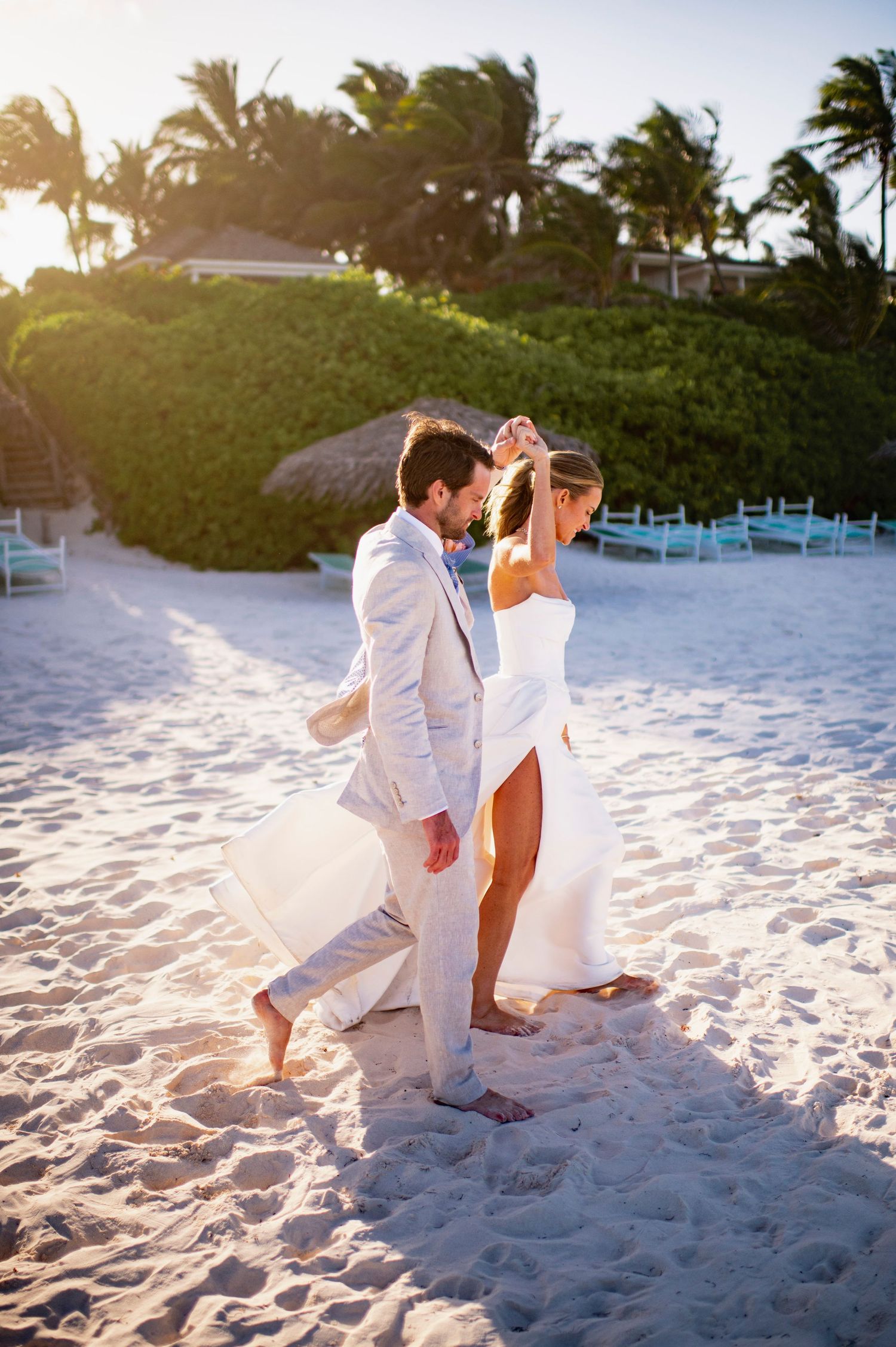 Couple dancing on beach at sunset with palm trees and beach chairs in background.