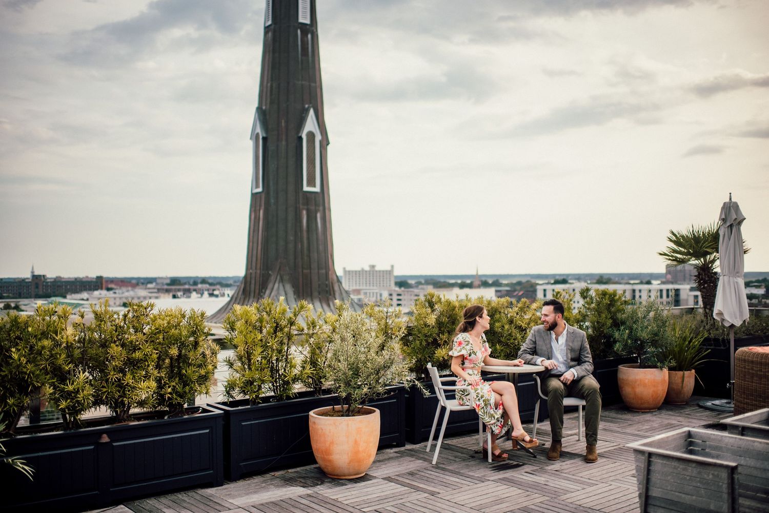 A panoramic view of a rooftop terrace with lounge chairs, potted plants and city skyline in the background.
