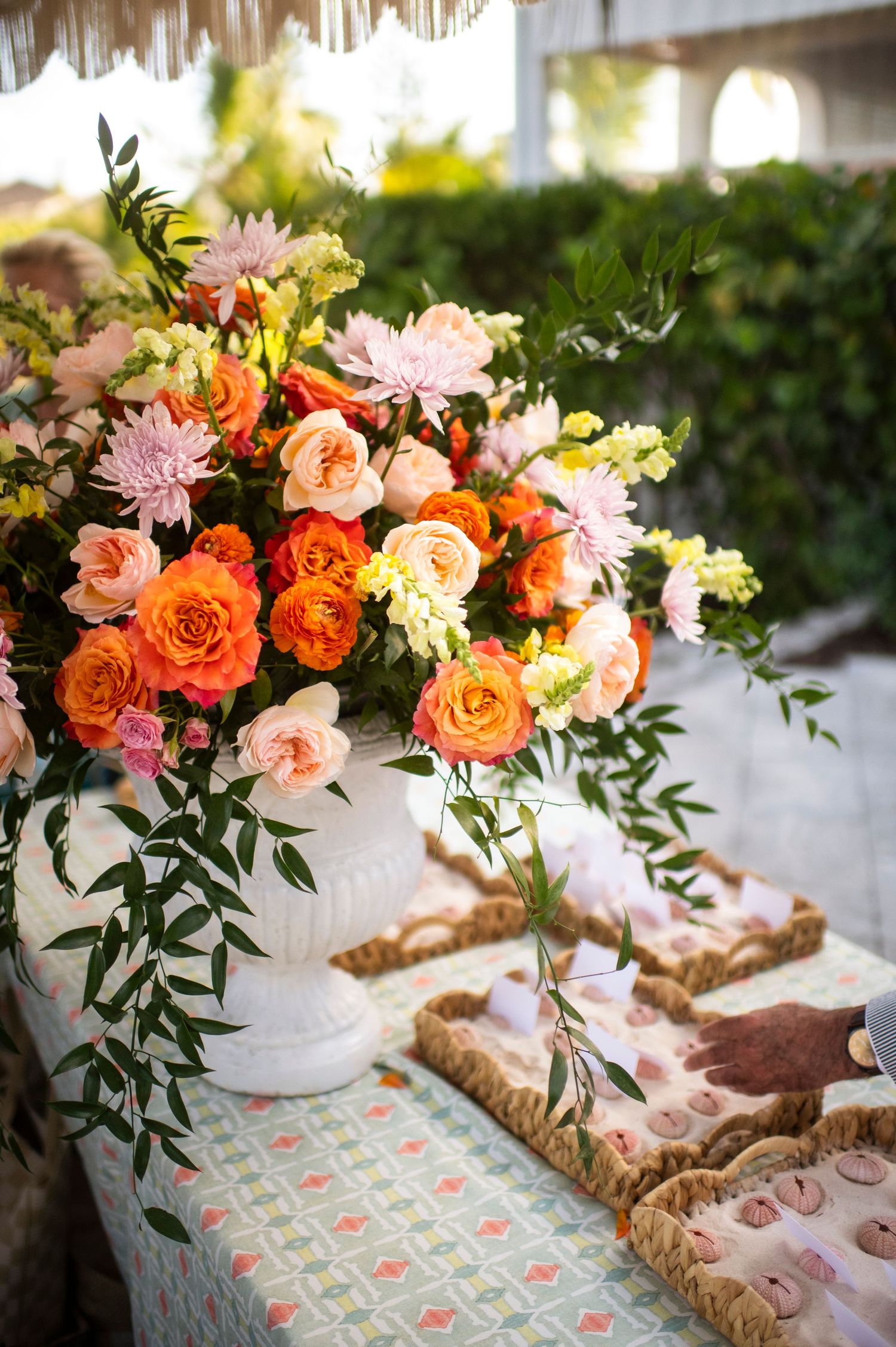 Vibrant floral arrangement with orange roses and pink blooms in a white vase on a patterned tablecloth.