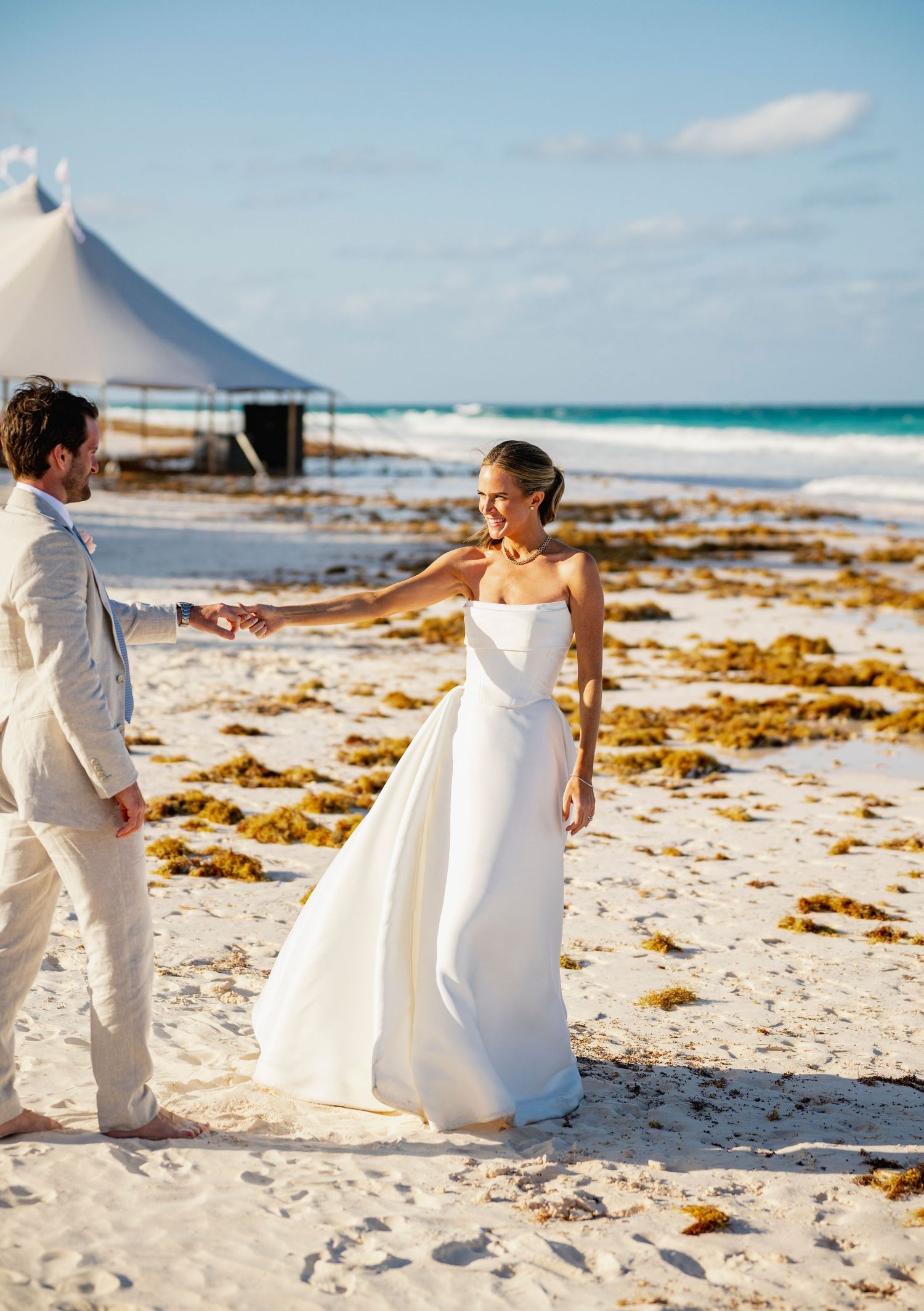Couple poses playfully on a scenic beach with white tents and seaweed-covered rocks in the background.