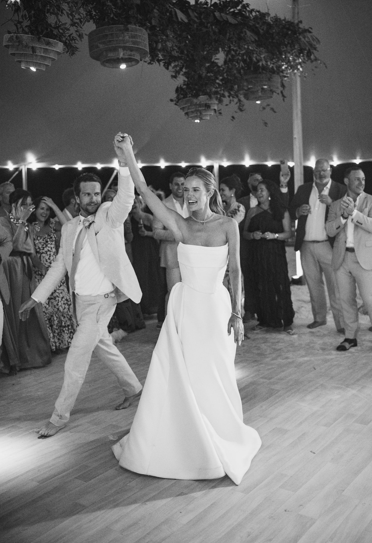 Black and white photo of wedding guests dancing on a wooden floor under string lights at an evening reception.
