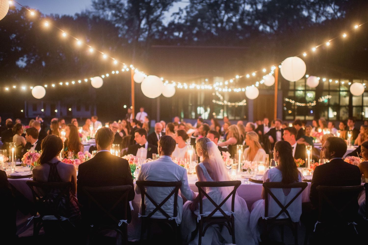 Outdoor evening wedding reception under string lights and paper lanterns with guests seated at long tables.