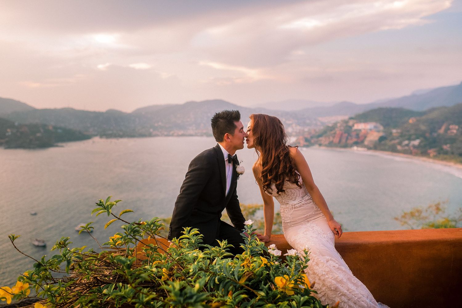 Romantic wedding couple embraces at sunset overlooking a scenic mountain lake with dramatic pink sky.