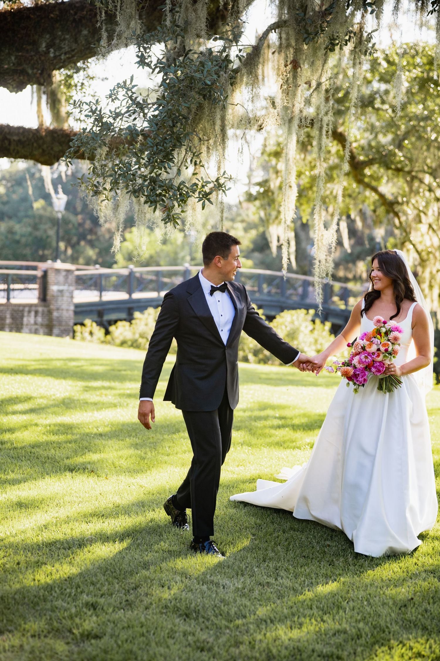 Newlywed couple walks on green lawn under Spanish moss at outdoor wedding venue on sunny day.