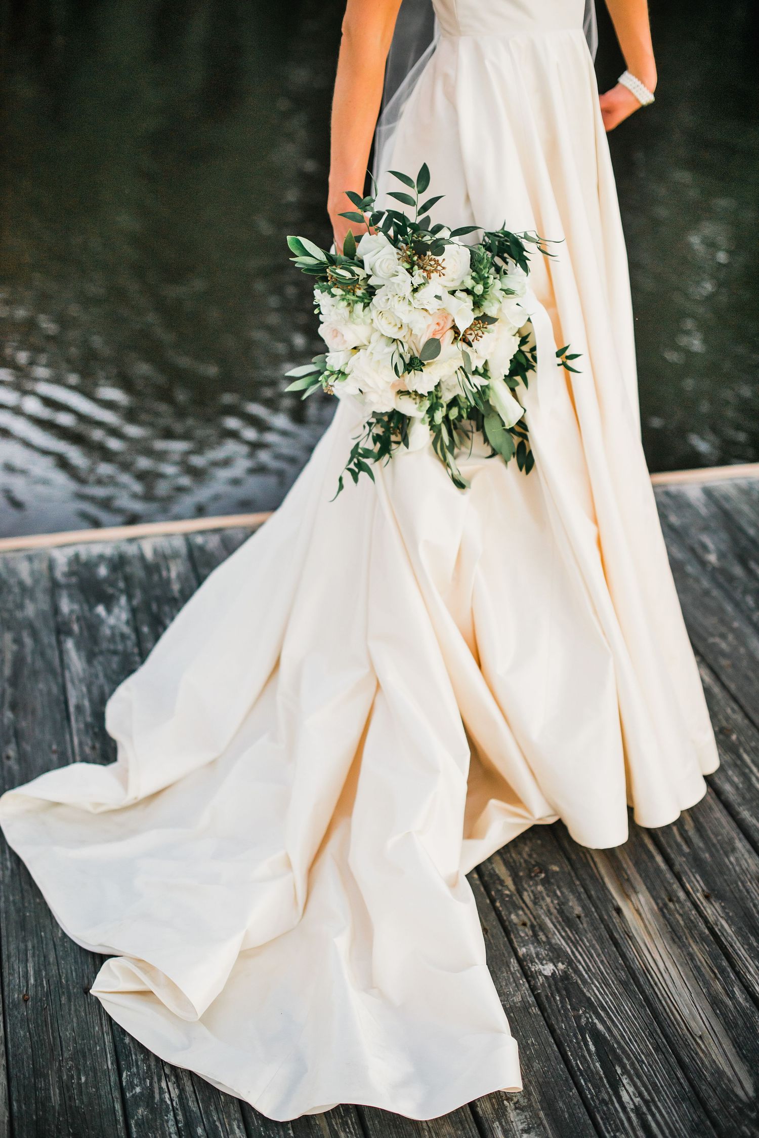 White wedding dress flowing elegantly over dark wooden deck with delicate floral bouquet featuring blush and green accents.