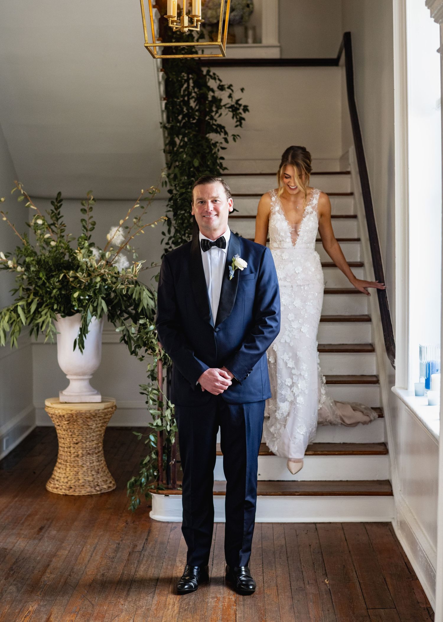 A couple shares a first look moment on a staircase with elegant floral arrangements and hardwood floors.