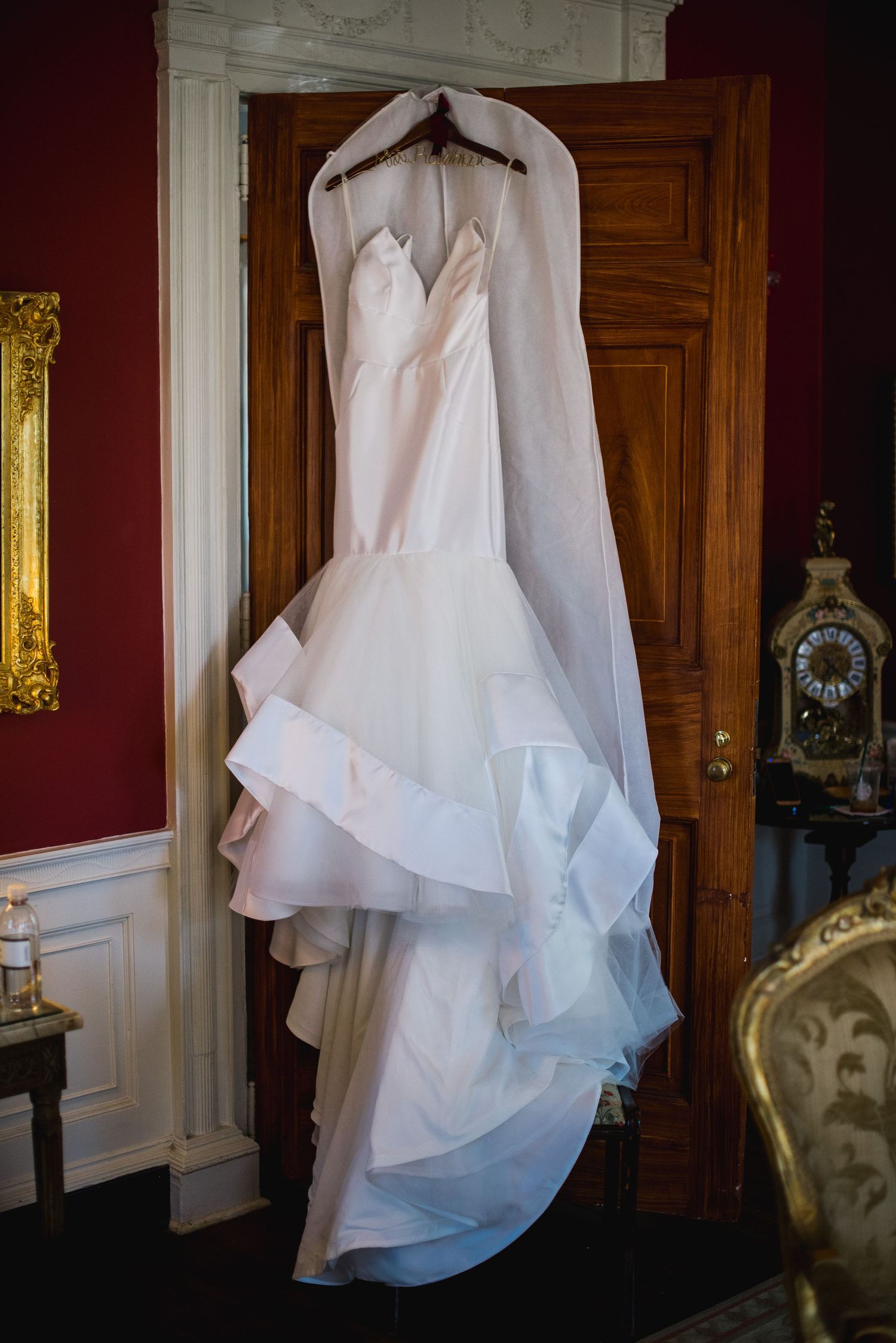 White wedding dress with ruffled skirt and plunging neckline hangs elegantly in a dark wood-paneled room.