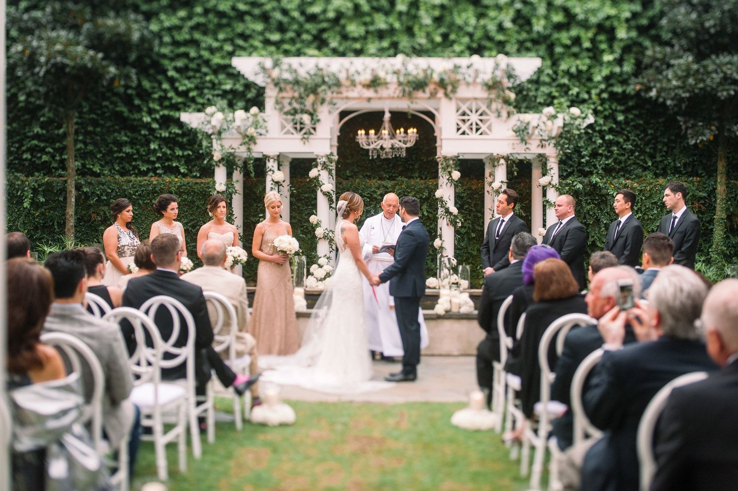 Outdoor garden wedding ceremony setup with white chairs and greenery along stone path.