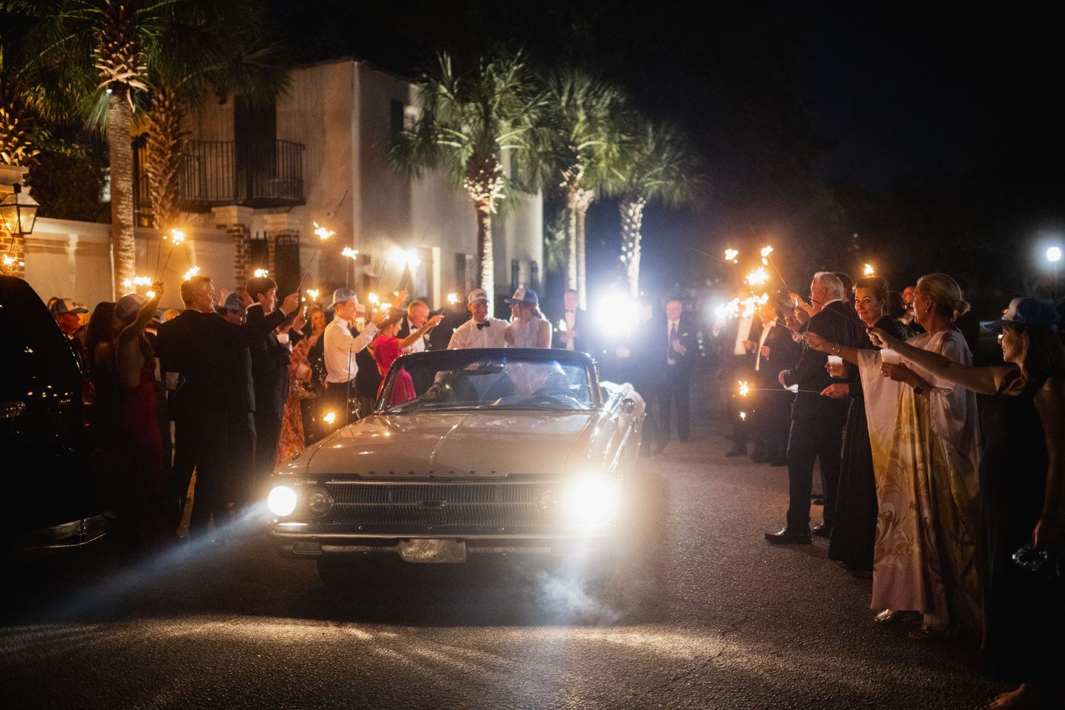 Cars with headlights illuminating a dark street at night with people gathered along the sides.