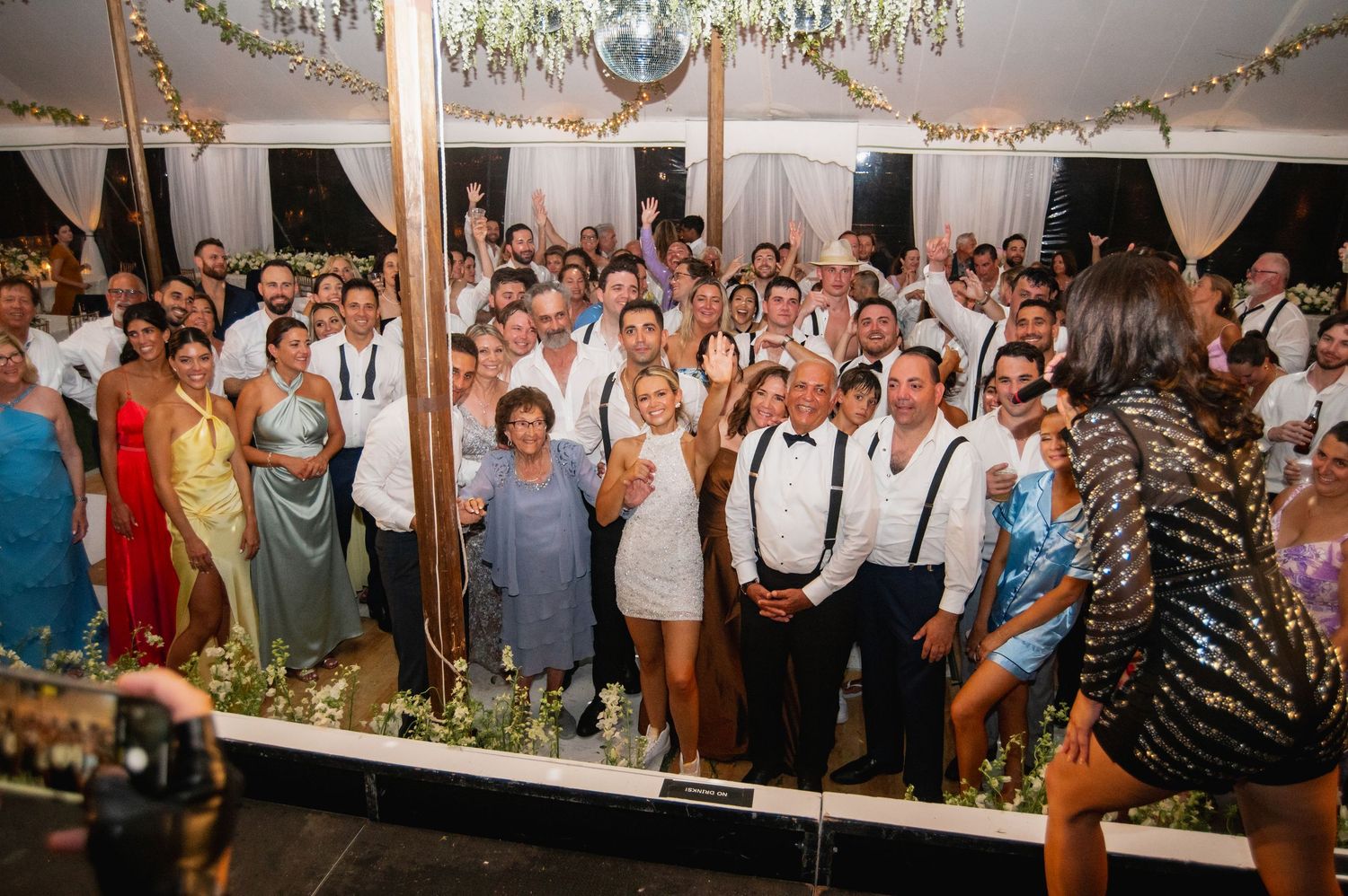 Large group of wedding guests gathered for celebration photo under chandelier and decorative lighting.