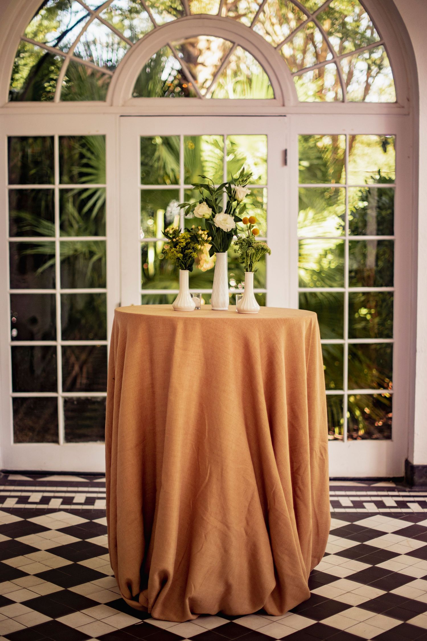 Elegant round cocktail tables with champagne-colored linens arranged in a room with arched windows and checkered floors.