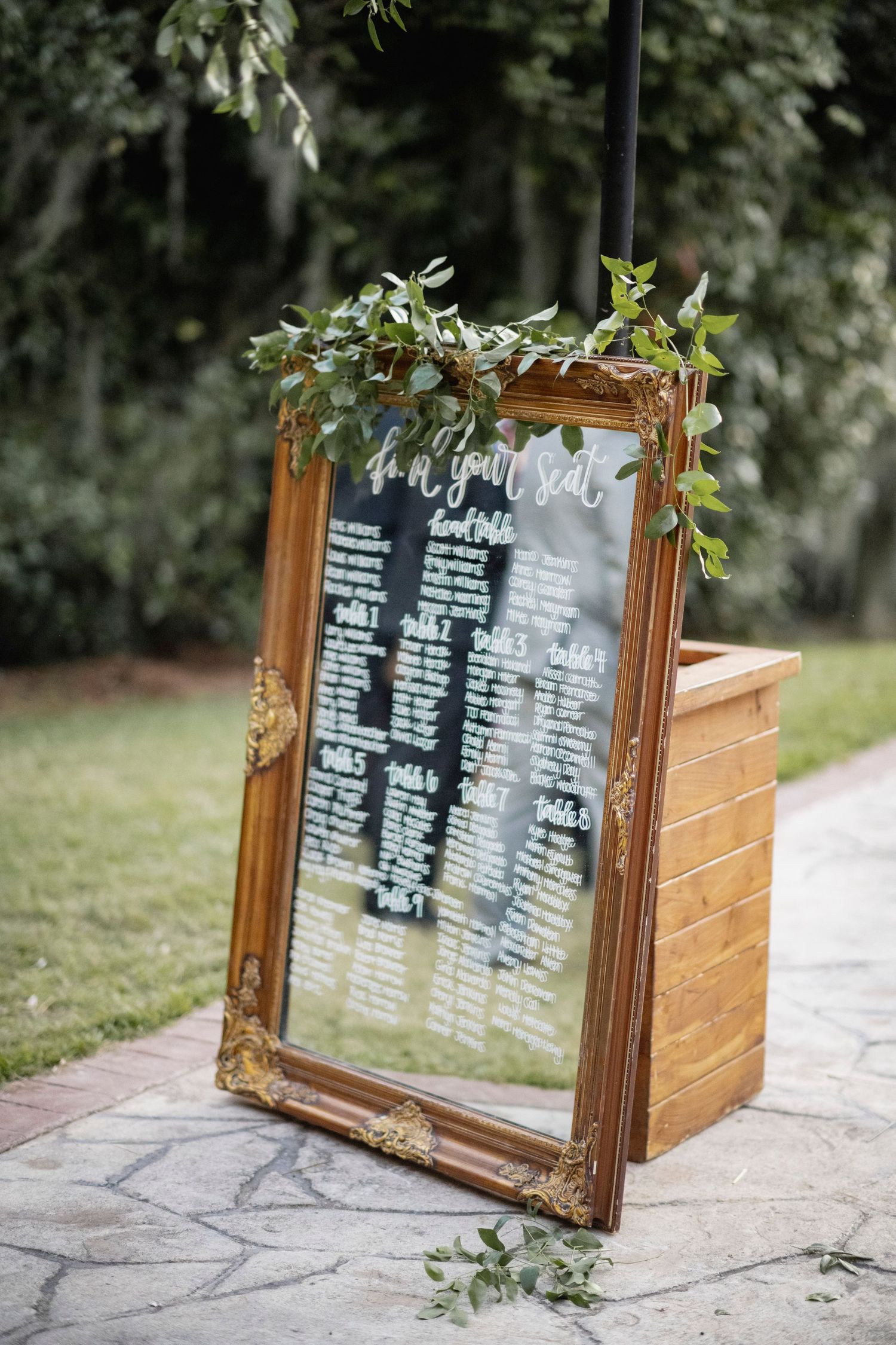 Elegant gold-framed mirror with handwritten seating chart and greenery decoration leans against wooden box at outdoor venue.