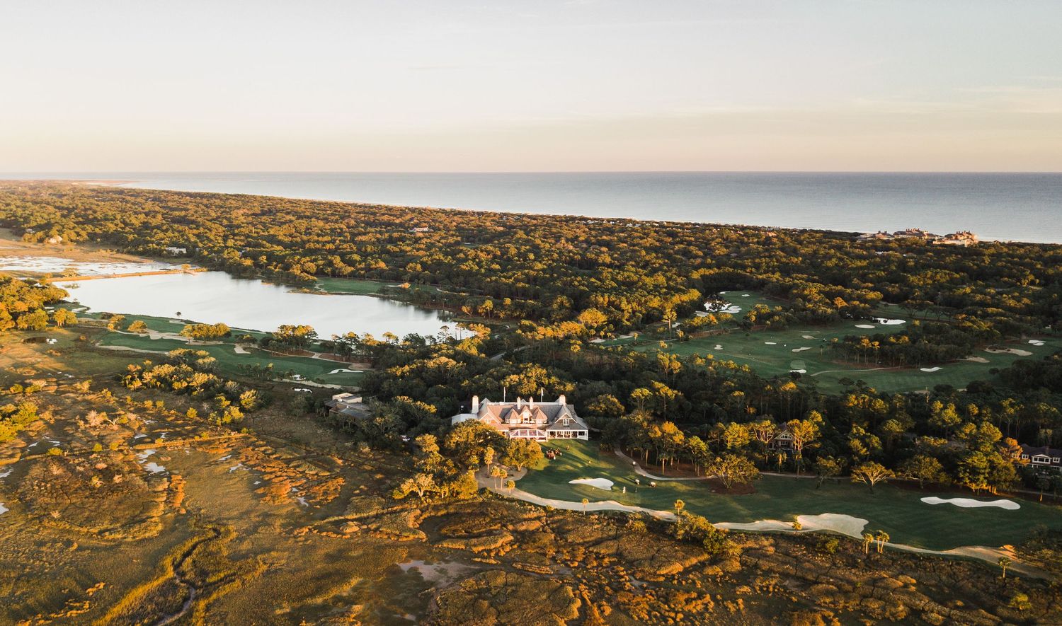 Aerial panoramic view of a coastal golf course with rolling hills and luxury homes at sunset.